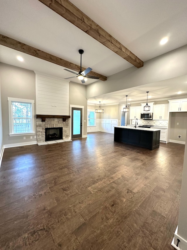 unfurnished living room featuring ceiling fan with notable chandelier, a stone fireplace, sink, beamed ceiling, and dark hardwood / wood-style flooring