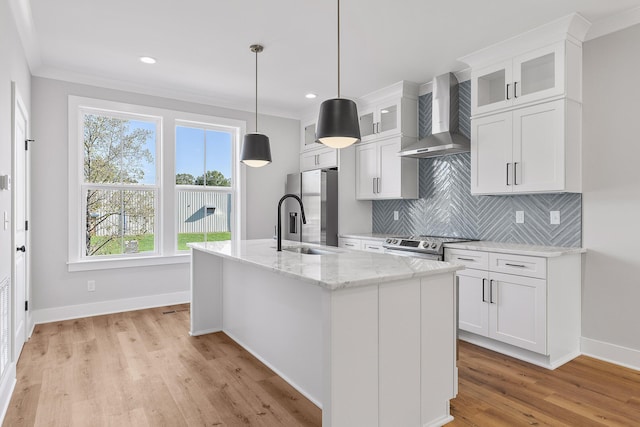 kitchen featuring wall chimney exhaust hood, stainless steel appliances, a kitchen island with sink, pendant lighting, and white cabinets