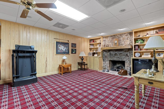 living room featuring carpet, a drop ceiling, a stone fireplace, ceiling fan, and built in features