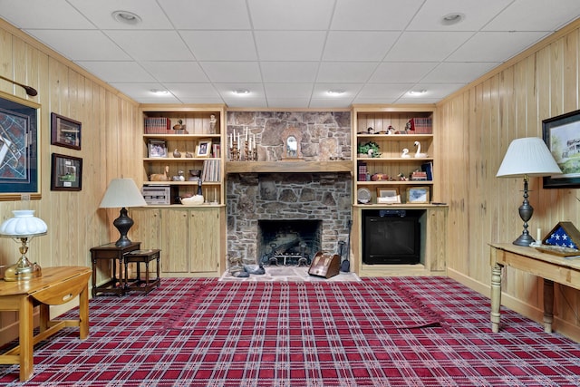 carpeted living room with built in shelves, wooden walls, and a stone fireplace