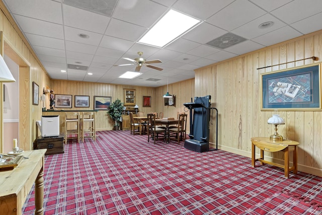 interior space featuring carpet flooring, ceiling fan, a drop ceiling, and wooden walls