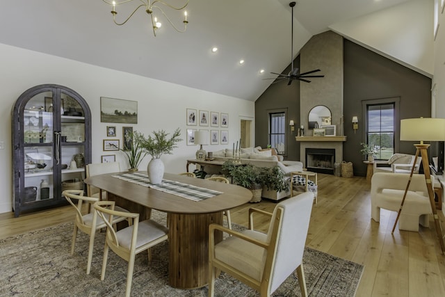 dining room with a large fireplace, light hardwood / wood-style flooring, high vaulted ceiling, and ceiling fan with notable chandelier