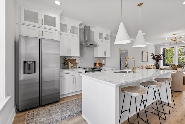 kitchen featuring white cabinetry, sink, stainless steel appliances, wall chimney range hood, and a center island with sink