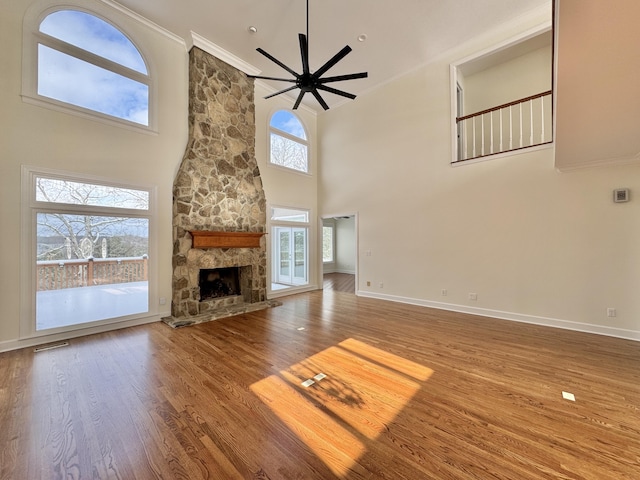 unfurnished living room featuring ceiling fan, a stone fireplace, crown molding, a towering ceiling, and wood-type flooring