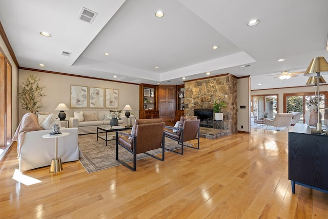 living room featuring a stone fireplace, crown molding, light wood-type flooring, a raised ceiling, and ceiling fan