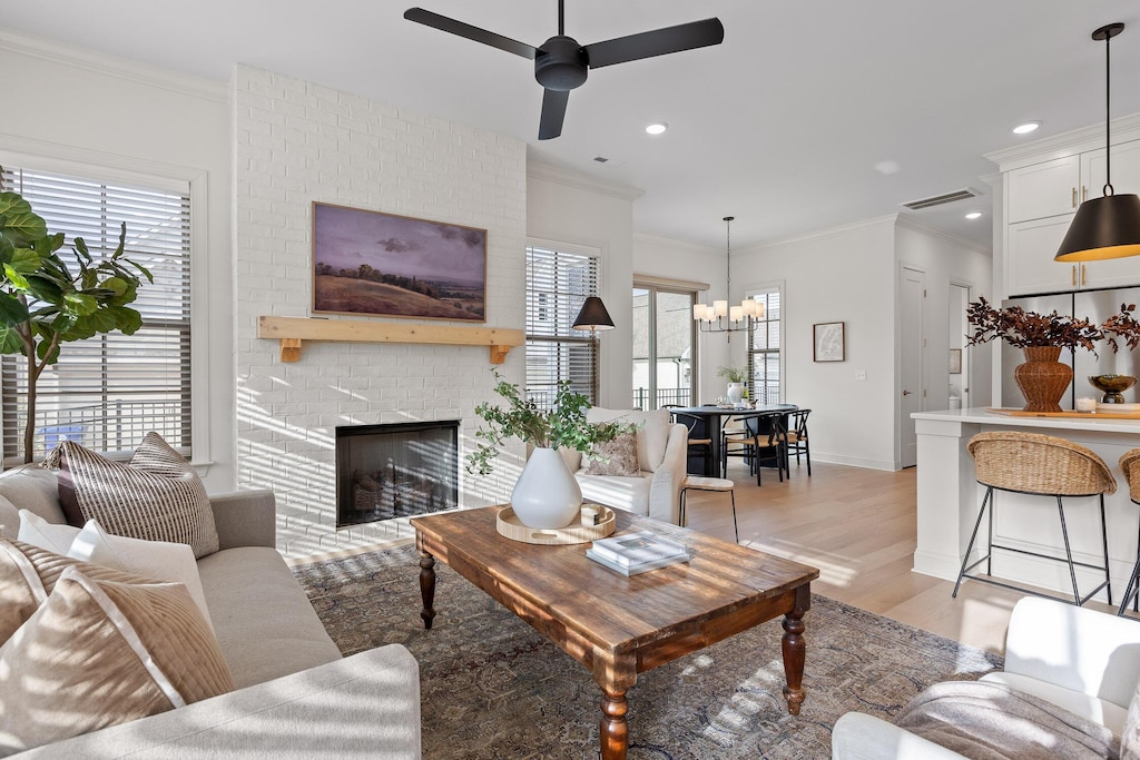 living room with light wood-type flooring, a brick fireplace, crown molding, and ceiling fan with notable chandelier