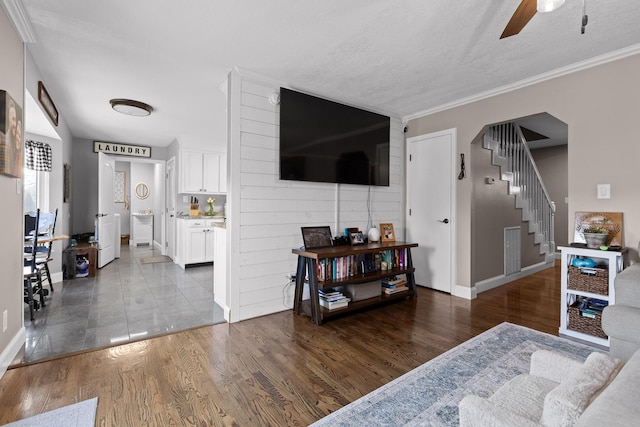 living area with stairway, a ceiling fan, baseboards, wood finished floors, and crown molding