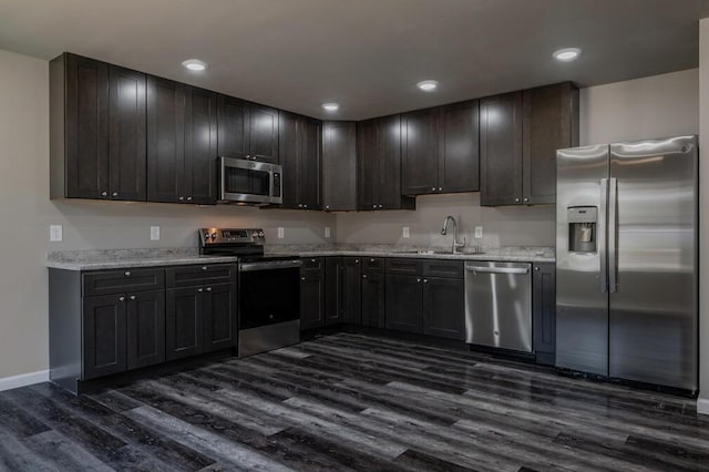 kitchen with a sink, baseboards, light stone countertops, appliances with stainless steel finishes, and dark wood-style flooring