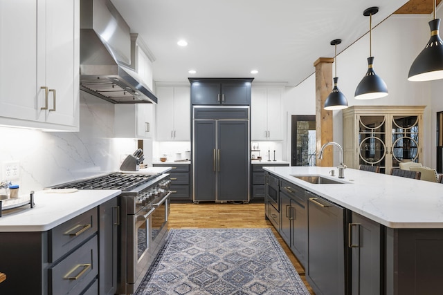 kitchen with wall chimney range hood, sink, premium appliances, white cabinets, and decorative light fixtures