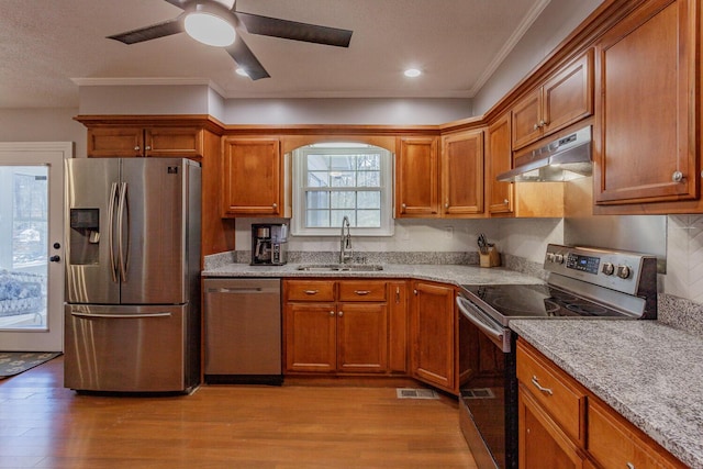 kitchen featuring light wood finished floors, under cabinet range hood, brown cabinets, appliances with stainless steel finishes, and a sink