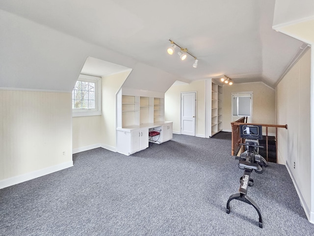 exercise area featuring lofted ceiling, rail lighting, dark colored carpet, built in desk, and built in shelves