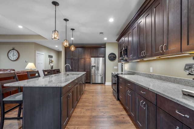 kitchen featuring appliances with stainless steel finishes, hanging light fixtures, a kitchen island with sink, light stone counters, and a breakfast bar area