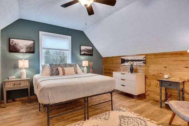 bedroom with vaulted ceiling, wood walls, light hardwood / wood-style floors, ceiling fan, and a textured ceiling