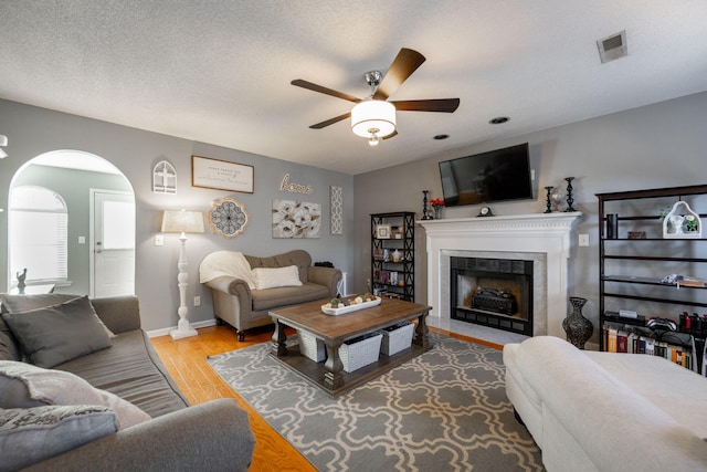 living room with a tiled fireplace, light hardwood / wood-style floors, a textured ceiling, and ceiling fan