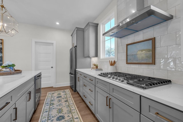 kitchen with wall chimney range hood, decorative light fixtures, backsplash, stainless steel appliances, and gray cabinets