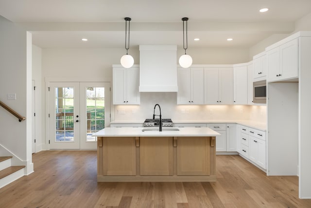 kitchen with a kitchen island with sink, white cabinetry, stainless steel microwave, custom range hood, and decorative light fixtures