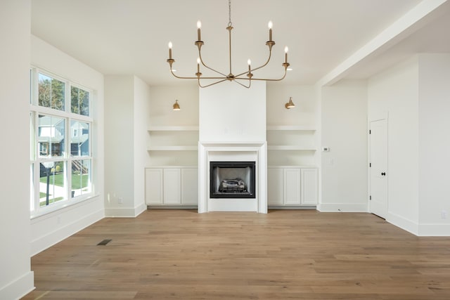 unfurnished living room featuring an inviting chandelier, a wealth of natural light, built in shelves, and hardwood / wood-style flooring