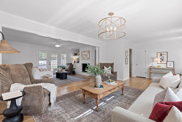 living room with ornamental molding, light hardwood / wood-style floors, a brick fireplace, ceiling fan with notable chandelier, and french doors