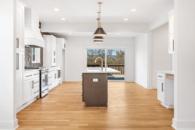 kitchen featuring pendant lighting, stainless steel stove, white cabinetry, an island with sink, and custom exhaust hood