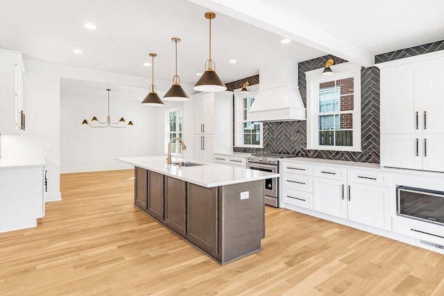 kitchen featuring decorative light fixtures, a center island with sink, stainless steel stove, custom range hood, and white cabinets