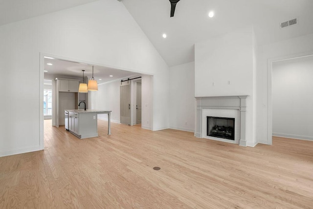 unfurnished living room featuring sink, ceiling fan, high vaulted ceiling, a barn door, and light wood-type flooring