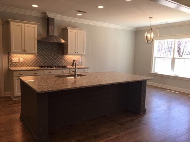 kitchen featuring white cabinetry, wall chimney range hood, light stone counters, and an island with sink