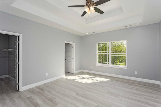 unfurnished bedroom featuring a spacious closet, light hardwood / wood-style flooring, a tray ceiling, a closet, and ceiling fan