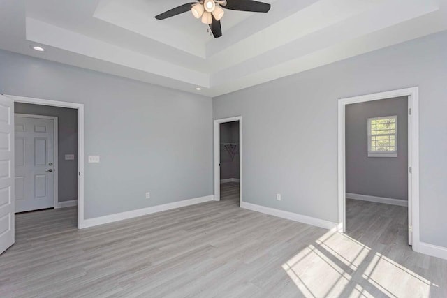 unfurnished bedroom featuring a walk in closet, a tray ceiling, a closet, ceiling fan, and light hardwood / wood-style floors