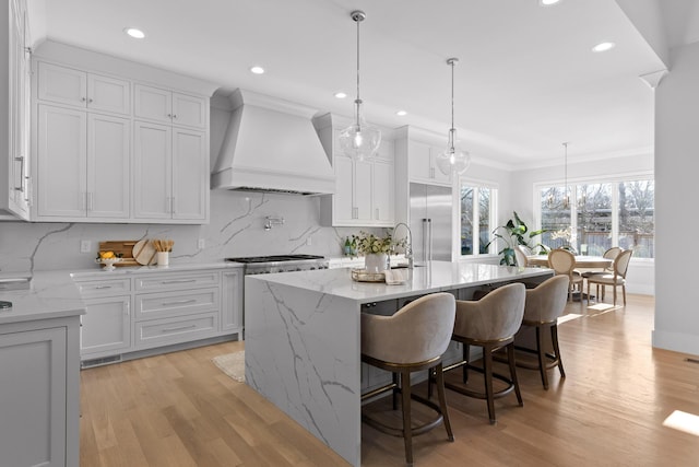 kitchen featuring light hardwood / wood-style flooring, a breakfast bar area, premium range hood, a center island with sink, and decorative light fixtures