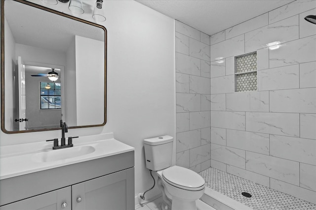 bathroom featuring ceiling fan, vanity, a tile shower, a textured ceiling, and toilet