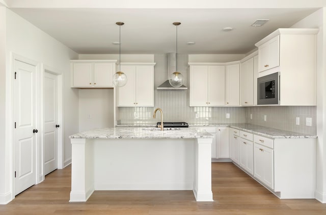 kitchen featuring an island with sink, wall chimney exhaust hood, light wood-style flooring, stainless steel microwave, and a sink