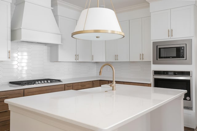 kitchen with custom exhaust hood, white cabinetry, decorative light fixtures, appliances with stainless steel finishes, and a kitchen island with sink