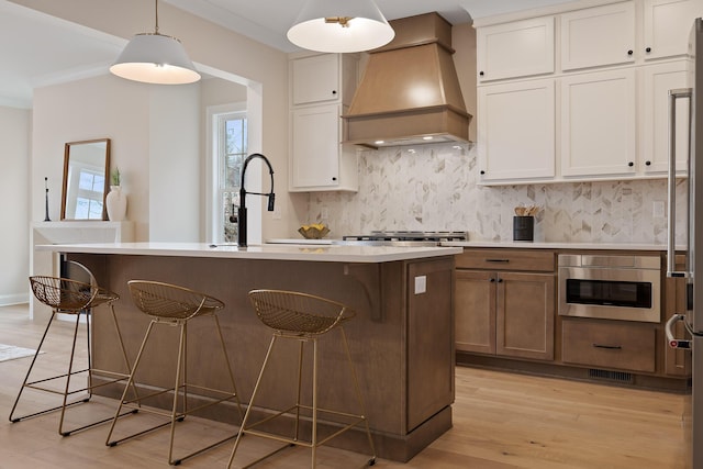 kitchen with a kitchen island with sink, hanging light fixtures, white cabinets, a kitchen bar, and custom exhaust hood