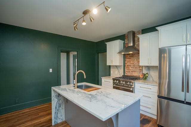 kitchen with sink, wall chimney range hood, an island with sink, stainless steel appliances, and white cabinets