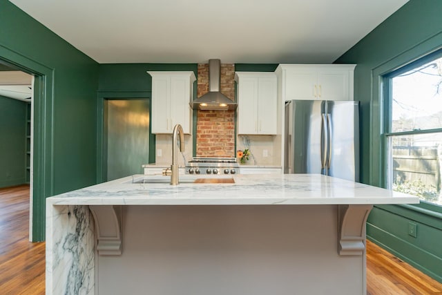 kitchen featuring stainless steel refrigerator, white cabinets, a center island with sink, and wall chimney range hood