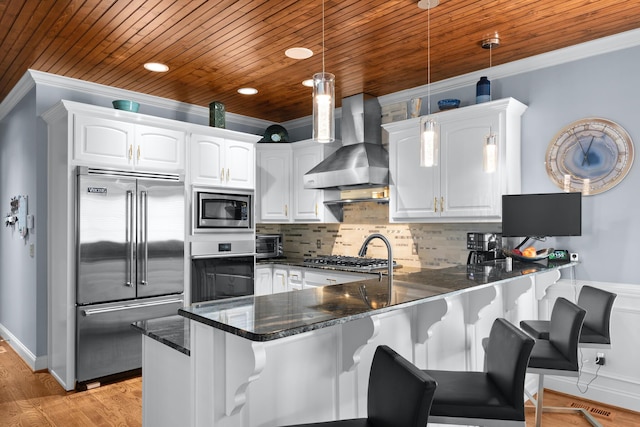 kitchen with white cabinetry, hanging light fixtures, built in appliances, kitchen peninsula, and wall chimney range hood