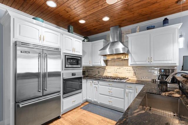 kitchen featuring wall chimney exhaust hood, sink, built in appliances, dark stone countertops, and white cabinets