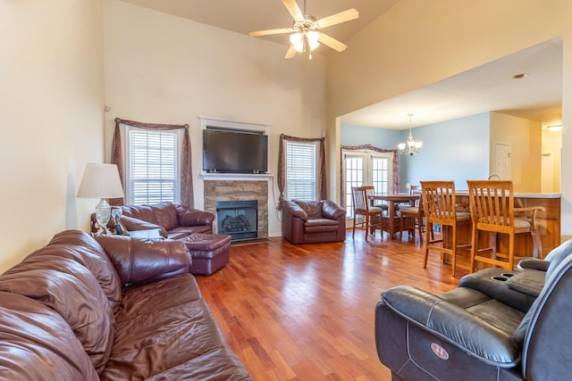 living room featuring hardwood / wood-style flooring, a towering ceiling, a stone fireplace, and ceiling fan with notable chandelier