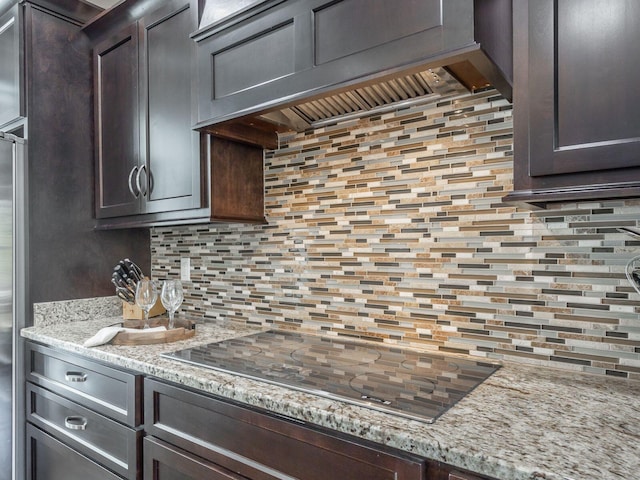 kitchen featuring dark brown cabinetry, light stone counters, tasteful backsplash, black electric cooktop, and custom exhaust hood