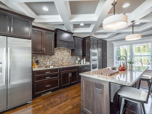 kitchen featuring premium range hood, light stone counters, pendant lighting, stainless steel appliances, and a kitchen island with sink