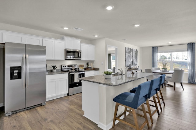 kitchen featuring appliances with stainless steel finishes, hardwood / wood-style floors, an island with sink, white cabinets, and dark stone counters