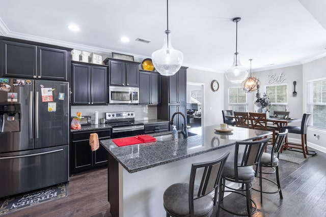 kitchen featuring hanging light fixtures, appliances with stainless steel finishes, a kitchen breakfast bar, dark stone counters, and a kitchen island with sink