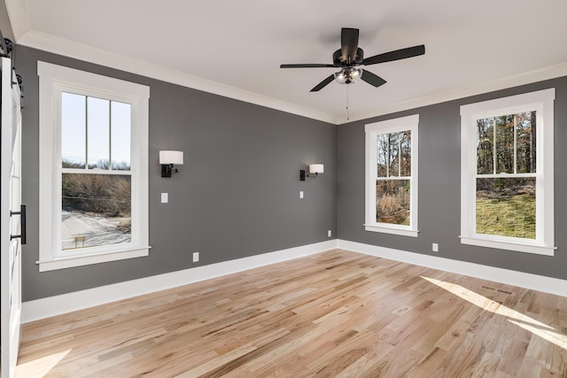 empty room featuring a barn door, ornamental molding, light wood-style floors, a ceiling fan, and baseboards