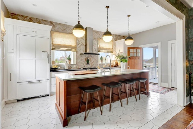 kitchen featuring white cabinets, a kitchen breakfast bar, hanging light fixtures, a kitchen island with sink, and wall chimney exhaust hood