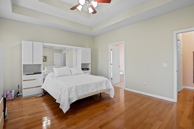 bedroom featuring a tray ceiling, ensuite bathroom, light wood-type flooring, and ceiling fan