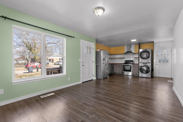 interior space with dark wood-type flooring, wall chimney exhaust hood, stacked washer / drying machine, stainless steel appliances, and backsplash