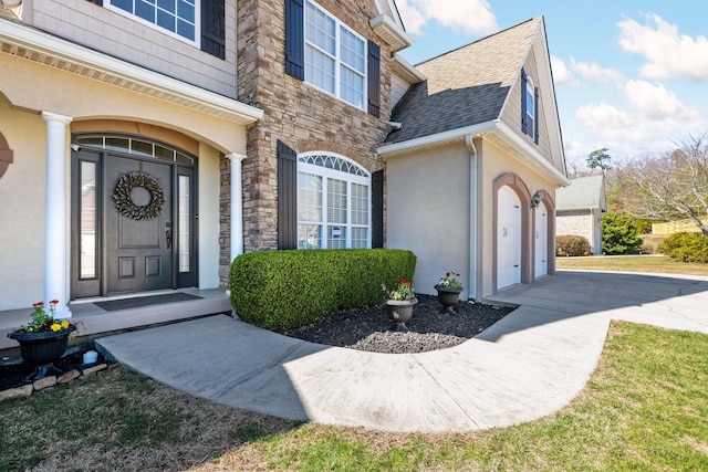 view of exterior entry with driveway, stone siding, roof with shingles, and stucco siding