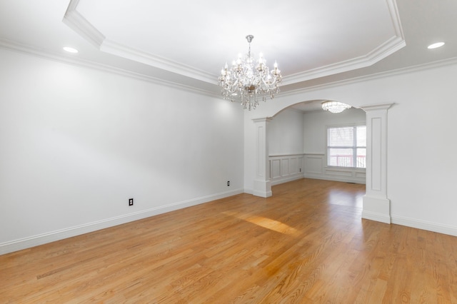 empty room with ornate columns, light wood-type flooring, ornamental molding, a tray ceiling, and a notable chandelier