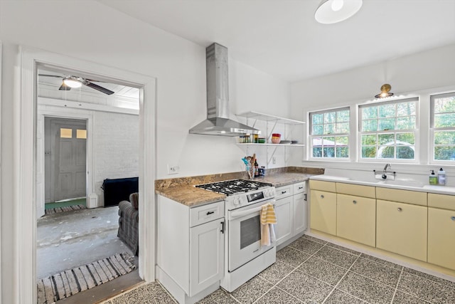 kitchen with wall chimney range hood, sink, white gas range oven, ceiling fan, and white cabinetry