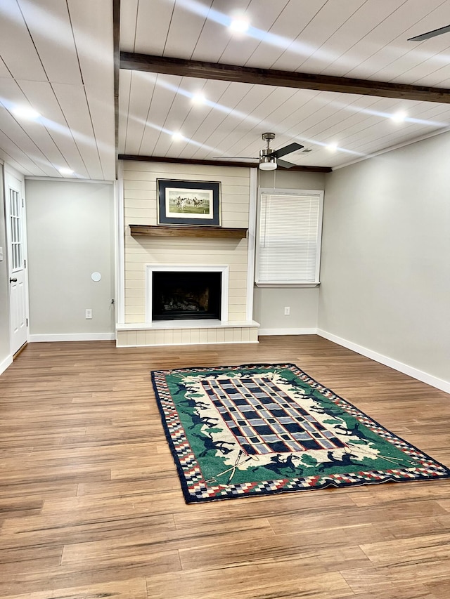 unfurnished living room with a fireplace, ceiling fan, wood finished floors, beamed ceiling, and baseboards